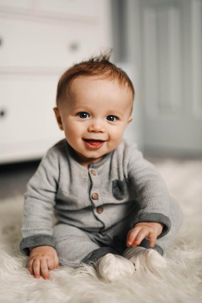 Smiling baby sitting on a soft white rug, wearing a cozy grey onesie. Can I Use Baby Shampoo Daily for gentle hair care? | babyblinkup.com
