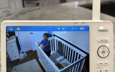 A VTech baby monitor displaying a live video feed of a young child standing inside a white crib. The monitor screen shows the room temperature, volume controls, and other settings. The background features a modern kitchen with a marble countertop. | How Does VTech Baby Monitor Work
